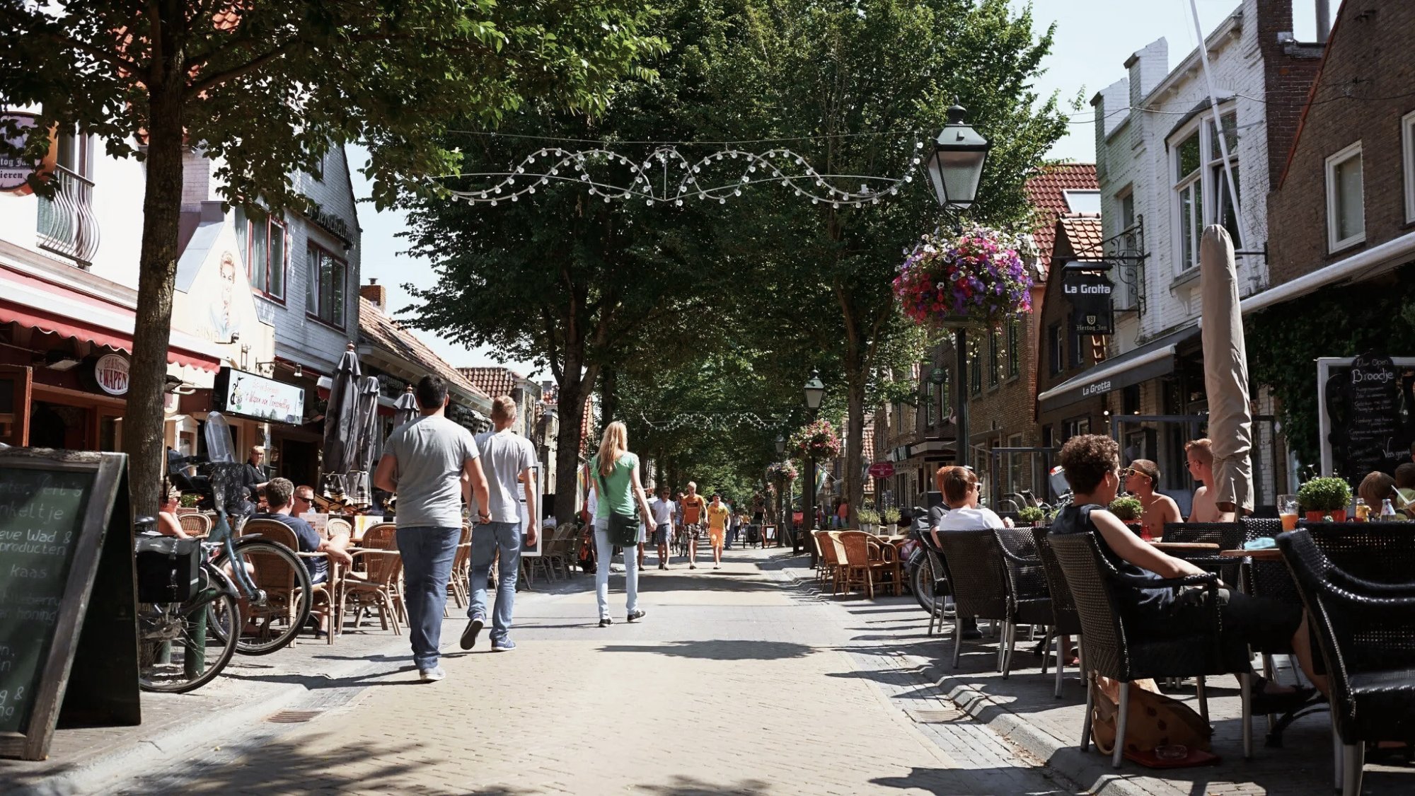 Zomers straatbeeld in Midsland op Terschelling met wandelaars, terrasjes en winkels onder groene bomen – levendig dorpscentrum op het Waddeneiland.