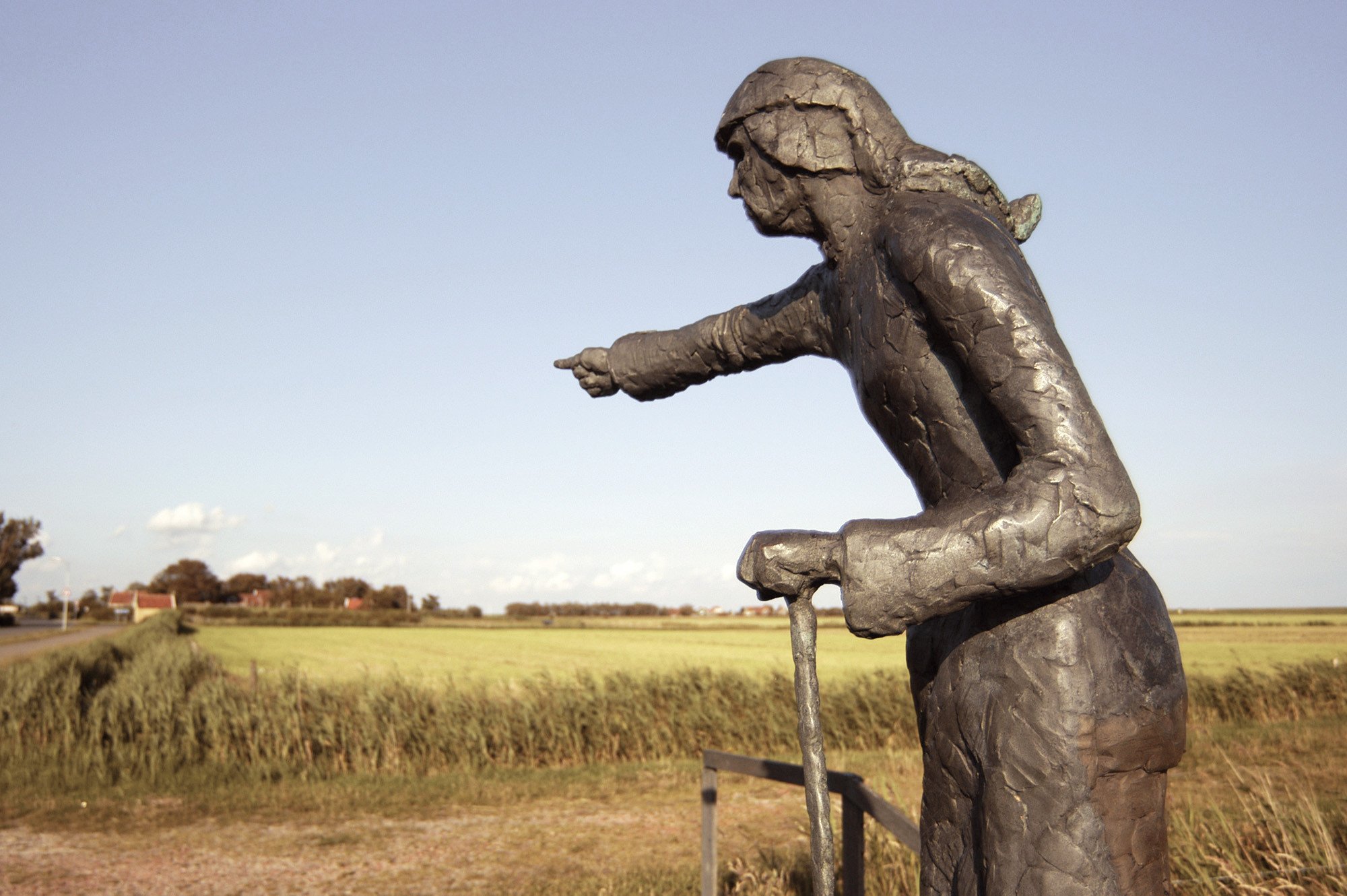 Beeld van de legendarische ‘Strieper Wyfke’ op Terschelling, een bronzen standbeeld van een vrouw die met uitgestrekte arm richting zee wijst, met op de achtergrond het open Friese landschap.
