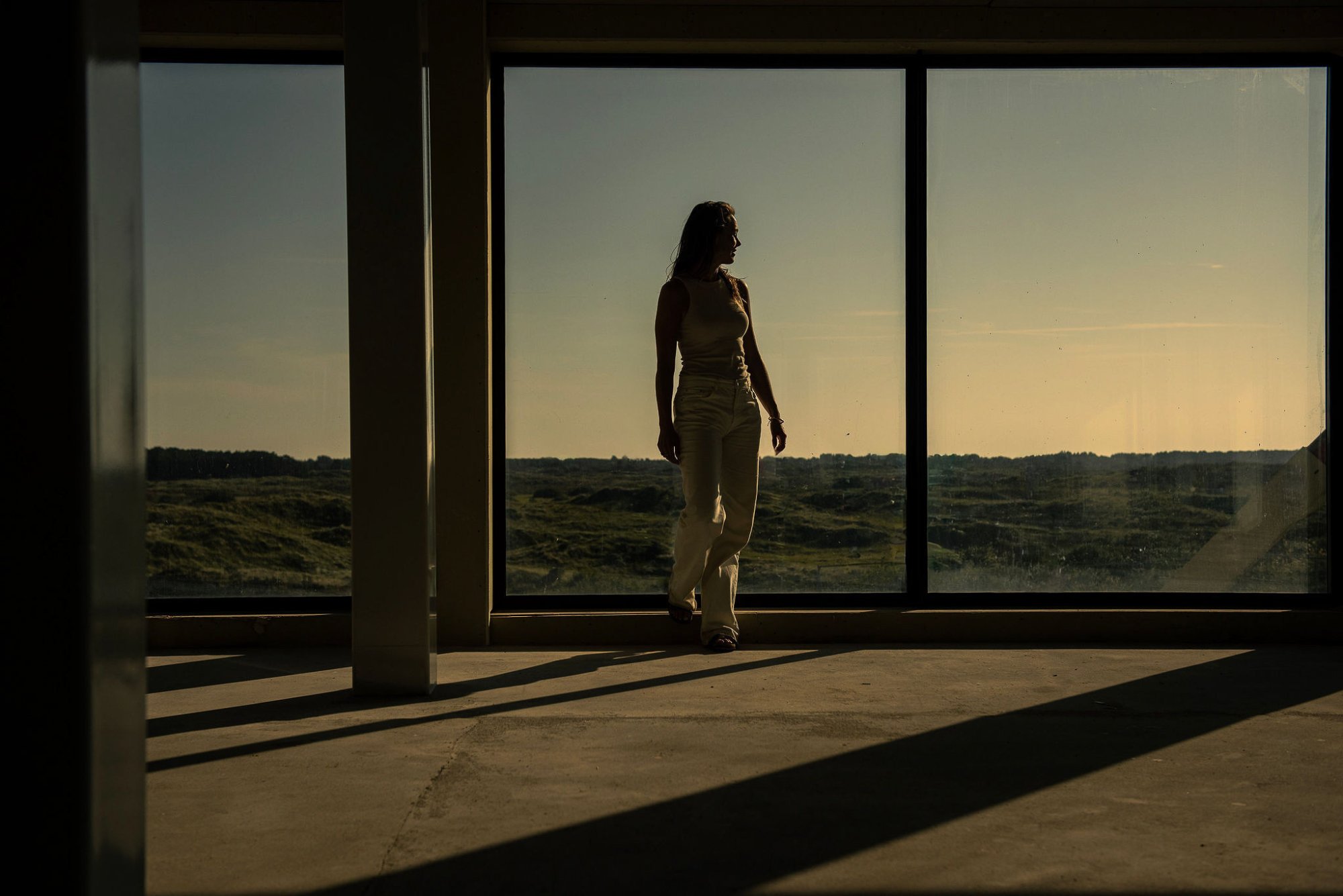 Een vrouw staat stil bij het grote raam van Elements Terschelling, omgeven door zacht avondlicht en uitzicht over de duinen — een moment van rust en reflectie.