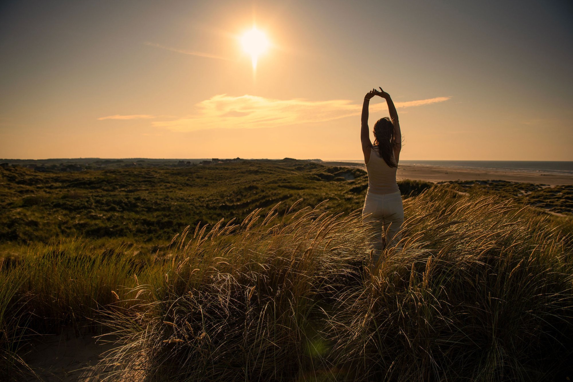 en vrouw strekt zich uit in het gouden avondlicht, omringd door wuivend duingras met uitzicht op zee — een moment van rust en verbinding met de natuur.