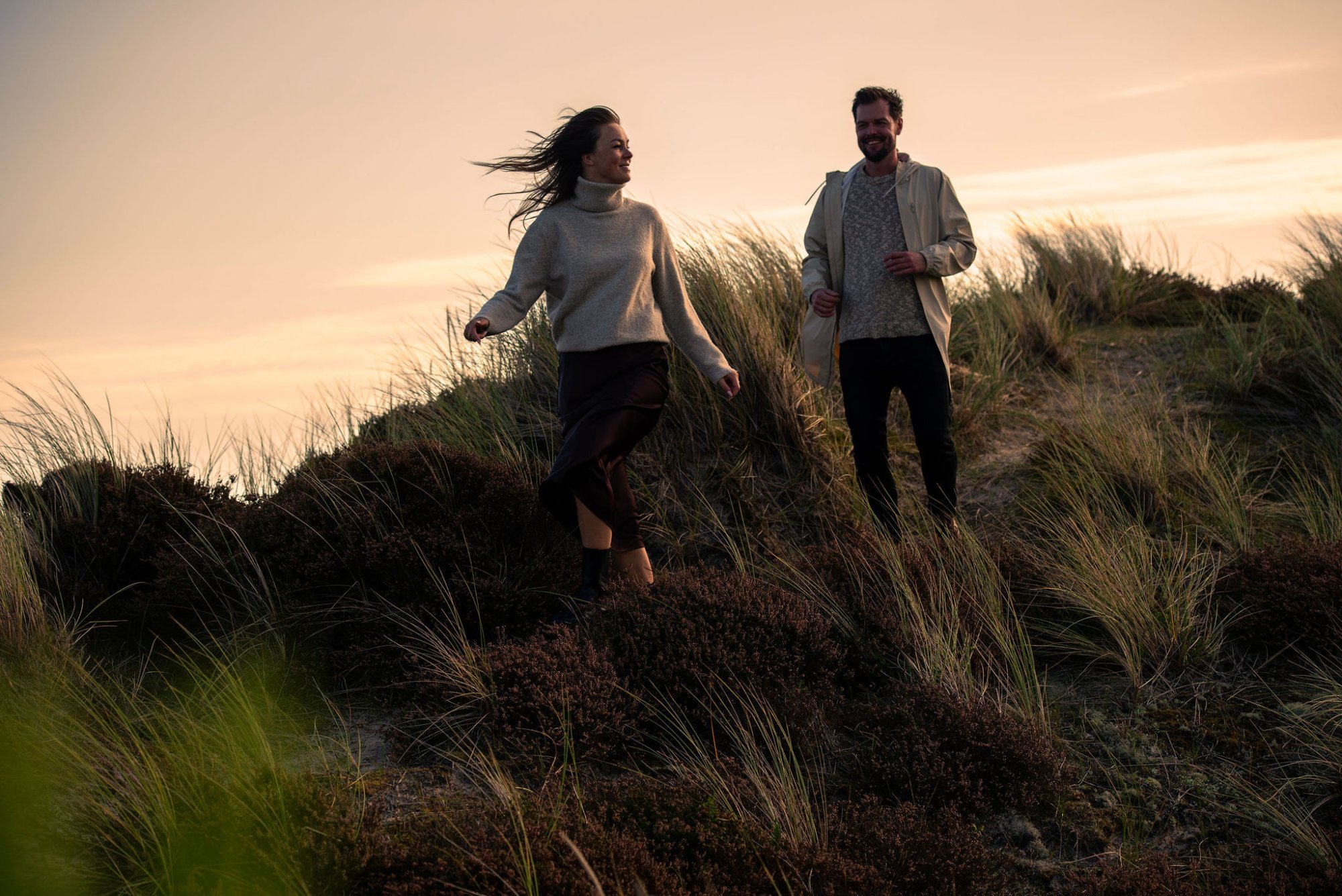 Twee mensen wandelen samen door de duinen van Terschelling bij zonsondergang, met wind in het haar en zacht avondlicht.