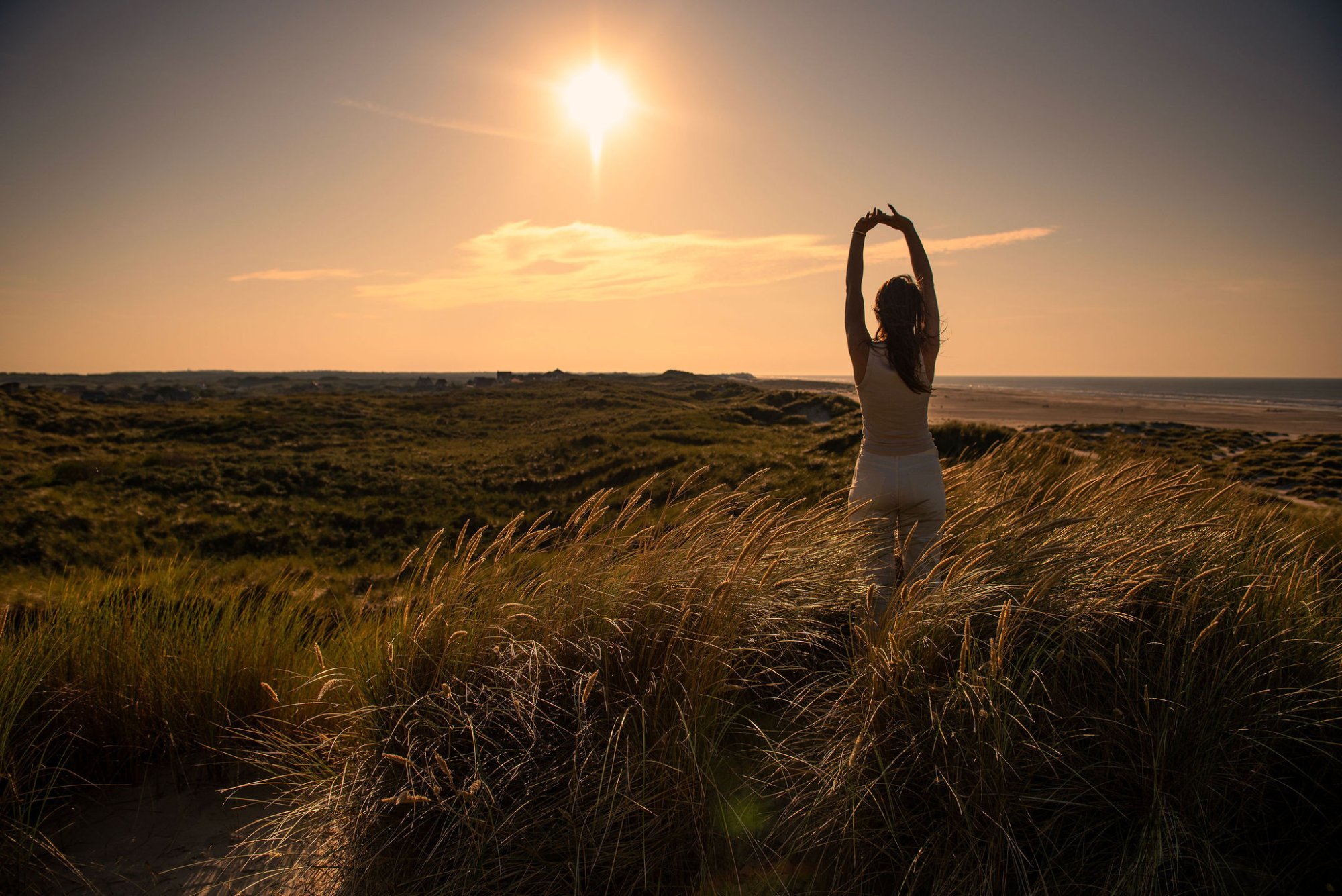 Een persoon doet yoga in de duinen van Terschelling, omringd door wuivend helmgras en warm avondlicht. De zon hangt laag boven de zee en vult de lucht met zachte goudtinten — een moment van stilte en verbinding met de natuur.