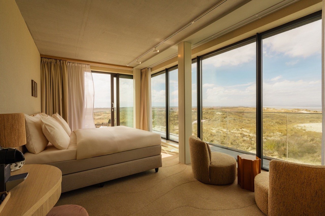 Deluxe hotelkamer bij Elements Terschelling met een tweepersoonsbed, twee fauteuils en ramen van vloer tot plafond met uitzicht over de duinen.