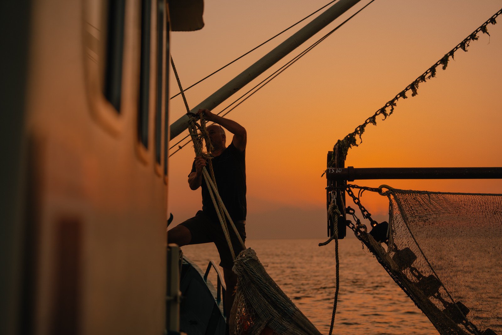 Een vissersboot op zee tijdens zonsondergang, met warme oranje gloed over het water. Een visser werkt aan de netten terwijl de laatste zonnestralen het schip en de golven zacht verlichten.