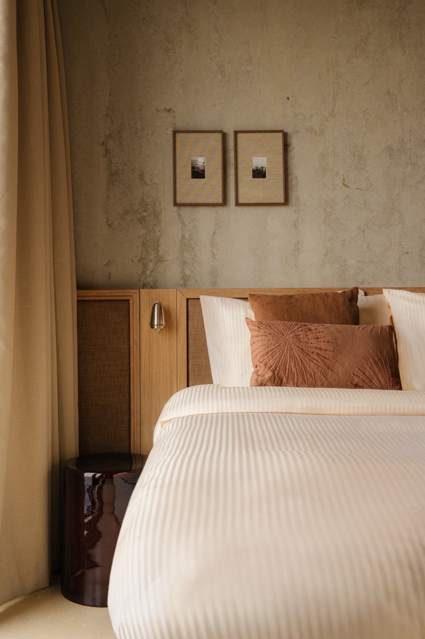 Tweepersoonsbed met wit gestreept beddengoed en terracotta kussens in een hotelkamer bij Elements Terschelling, met een houten hoofdbord, leeslampje en twee ingelijste foto's aan de muur.
