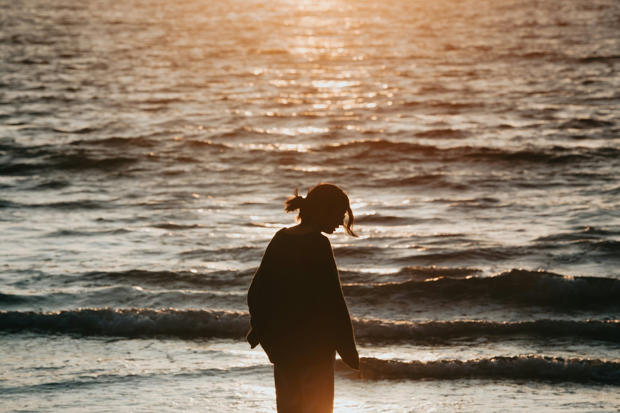 Silhouet van een vrouw aan de vloedlijn, met de zee op de achtergrond in het laatste zonlicht.