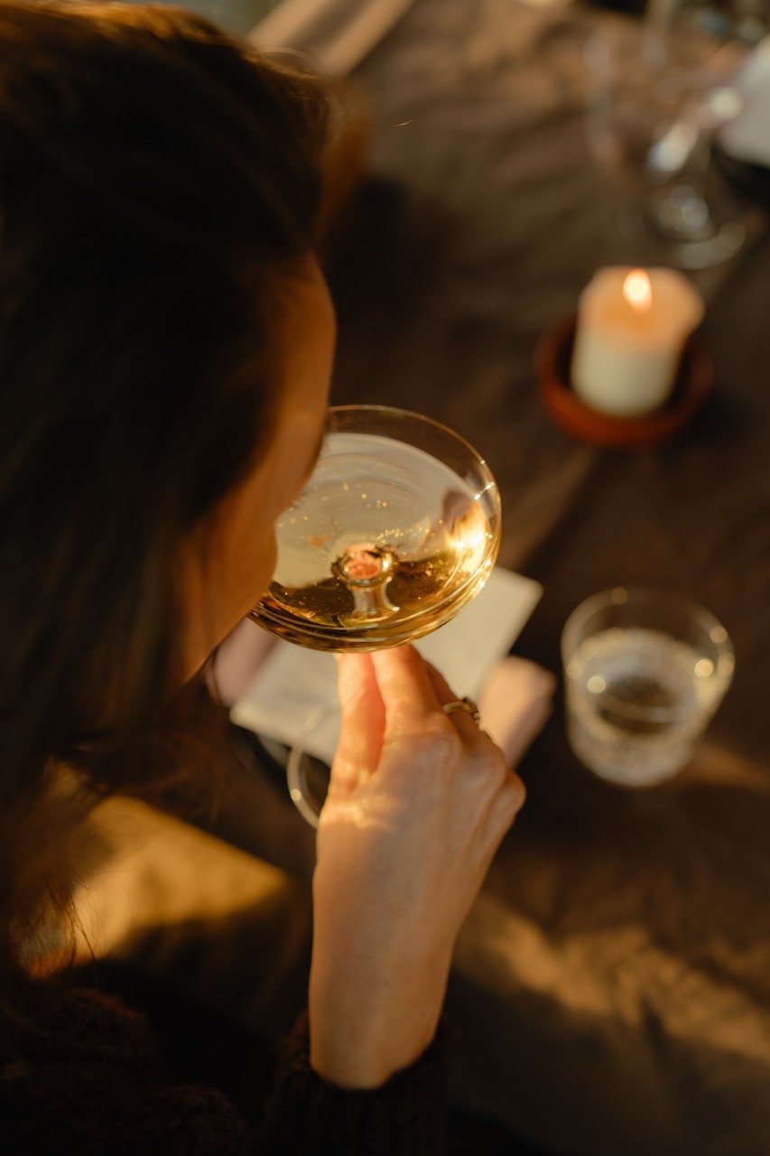 Vrouw drinkt uit een coupeglas aan tafel in restaurant Dún bij Elements Terschelling, met een brandende kaars en een glas water op tafel.