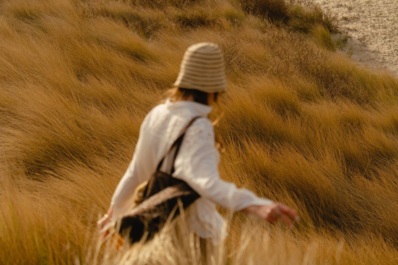 Vrouw met een gestreepte hoed en een tas over haar schouder loopt door het helmgras in de duinen bij Elements Terschelling.