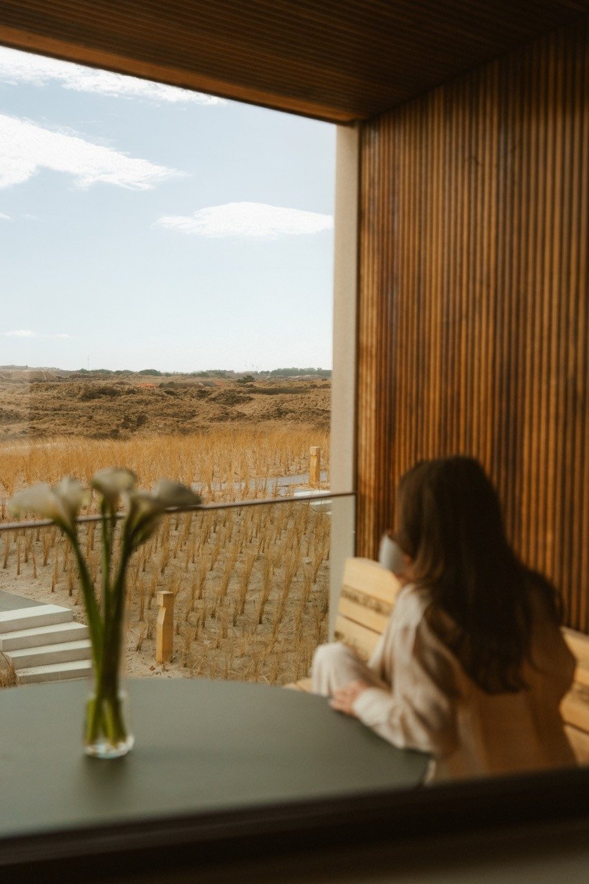 Vrouw zit met een kop koffie op het terras van het Royale Duinzicht-appartement bij Elements Terschelling en kijkt uit over de duinen, met calla's in een vaas op tafel.
