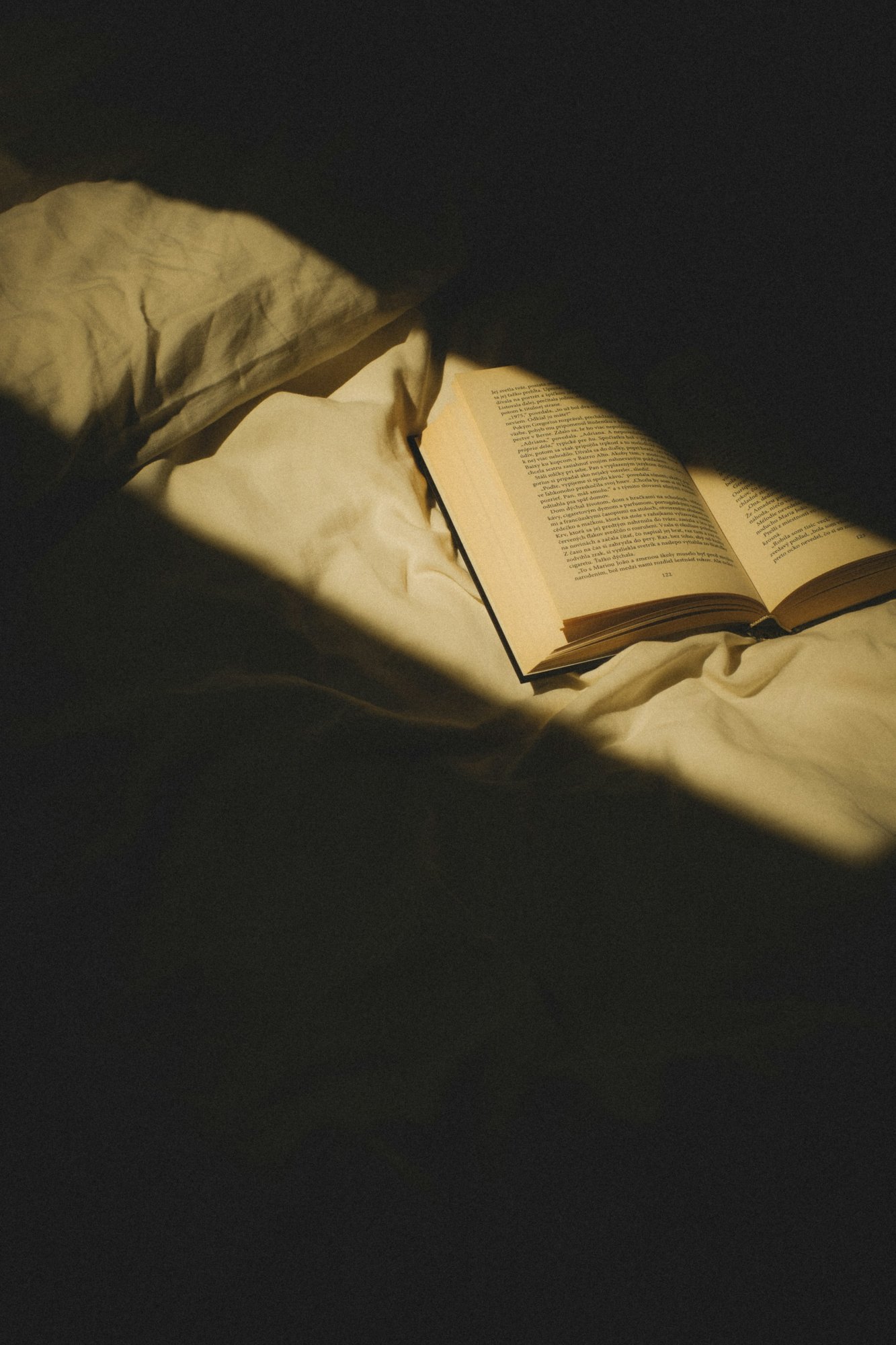 Opengeslagen boek op een onopgemaakt bed met wit beddengoed, in een streep zonlicht in een verder donkere kamer.