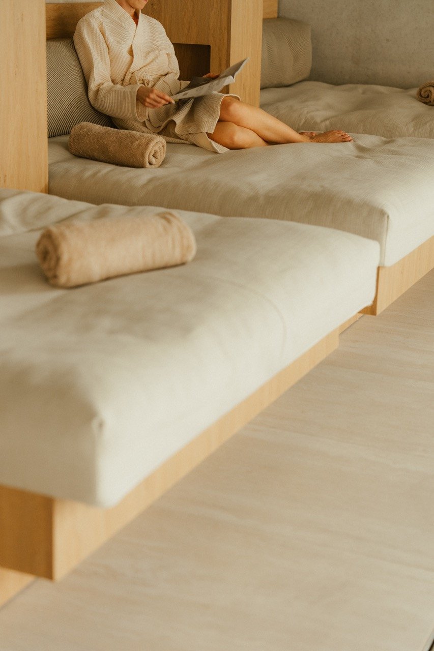Vrouw in een badjas leest een tijdschrift op een ligbed in de rustruimte van de wellness bij Elements Terschelling, met opgerolde handdoeken op de bedden.
