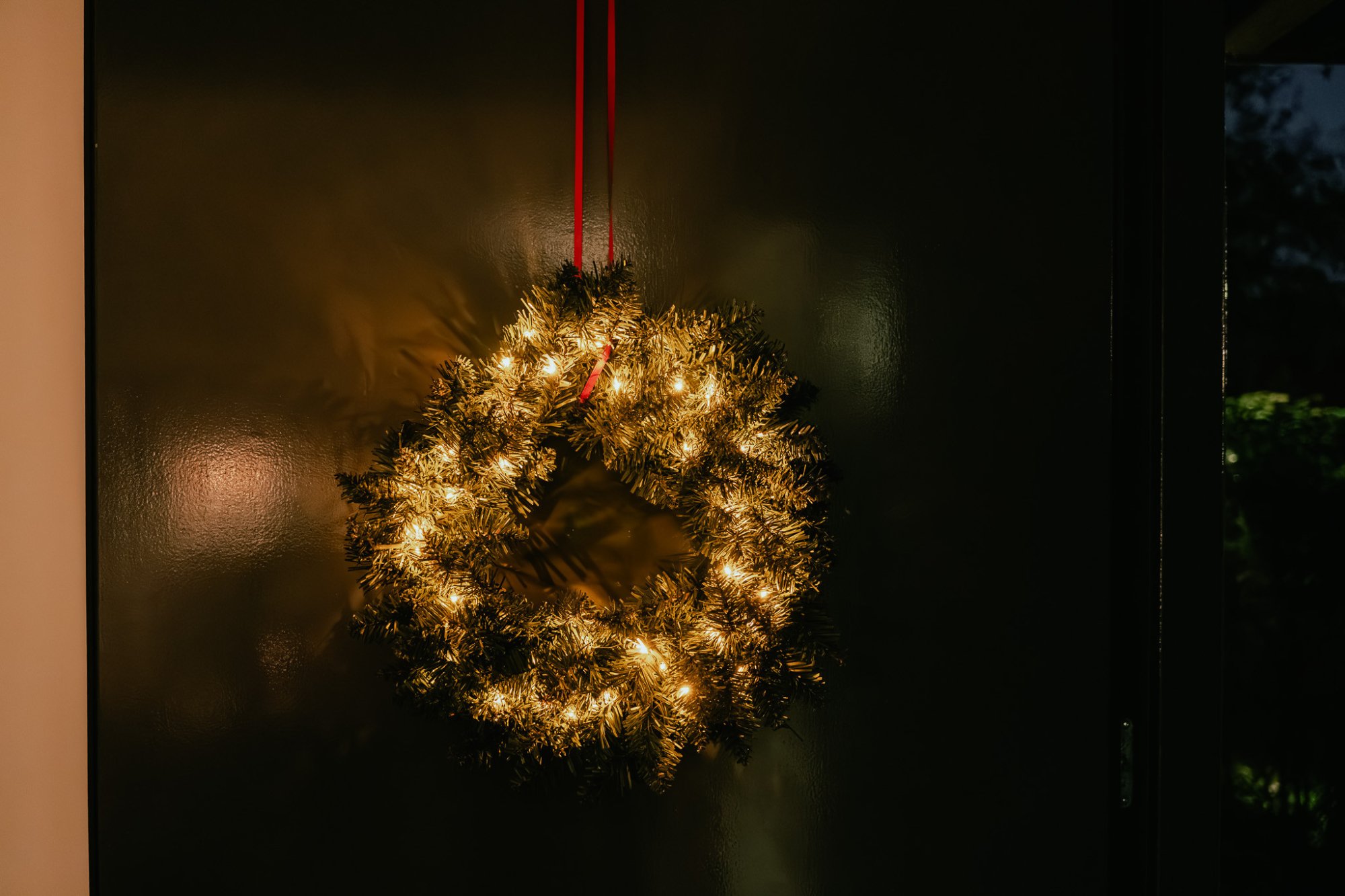 Verlichte kerstkrans met warme lichtjes aan een donkere voordeur – een sfeervolle welkom bij aankomst in het vakantiehuis.