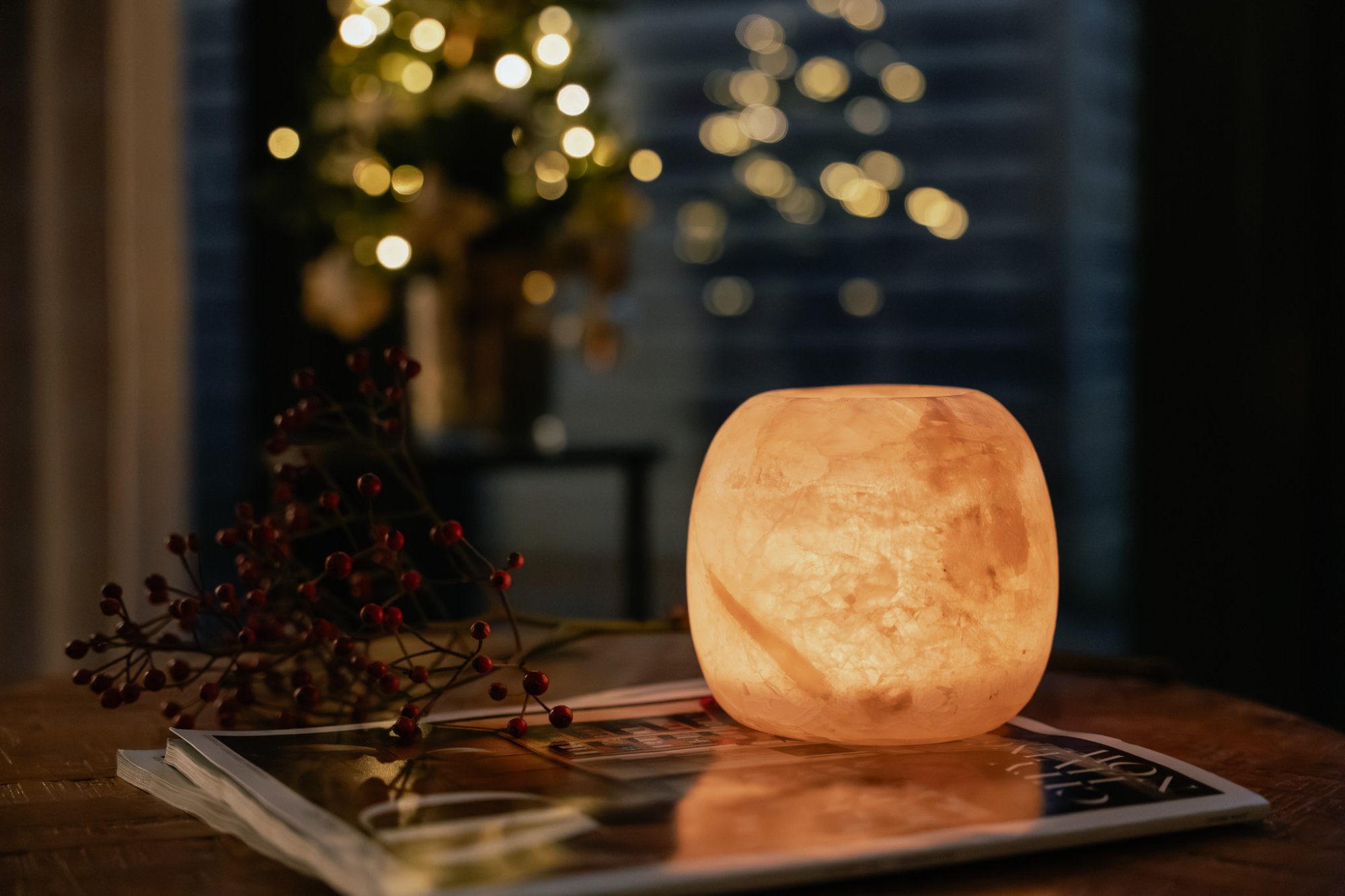 Zacht verlichte waxinelichthouder op een houten tafel met rode besjes en magazines, op de achtergrond kerstlichtjes – een warm welkom in winterse sferen.
