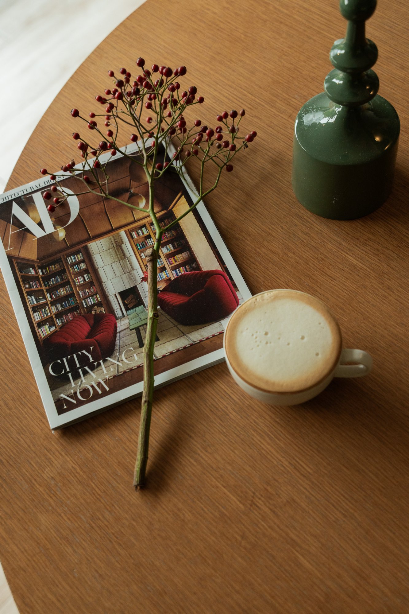Kopje koffie op een houten tafel met een tijdschrift, bloemen en een groene kandelaar.